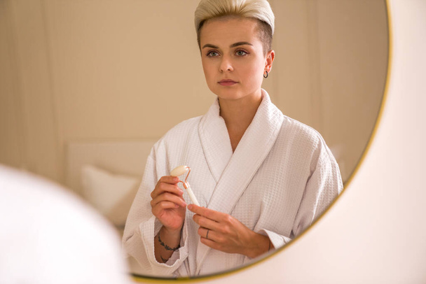 Portrait of serious caucasian female looking at her reflection at the mirror and using jade roller for skin treatment at bathroom - Foto, Imagem