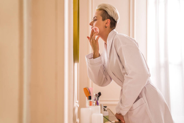 Pretty blonde young female wearing white bathrobe using cotton pads while remove make up from her face. Beauty routine concept - 写真・画像