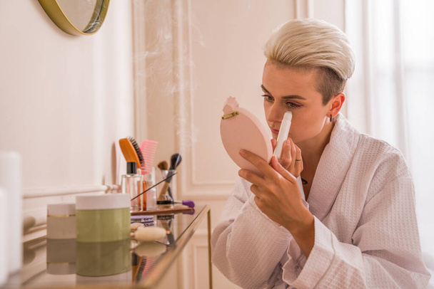 Enjoying the moment. Waist up portrait view of the short haired woman looking at the hand mirror while applying roller gel under her eyes - Фото, изображение