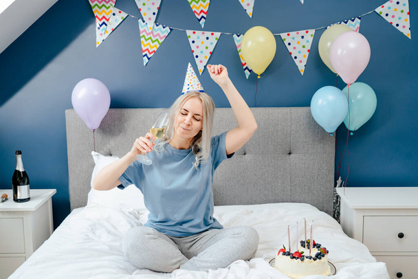 Happy emotional woman in pajama and party cap dancing on the bed with champagne glass in the hand and festive cake on the bed. Time for yourself Celebration at home. Happy birthday concept - Fotó, kép