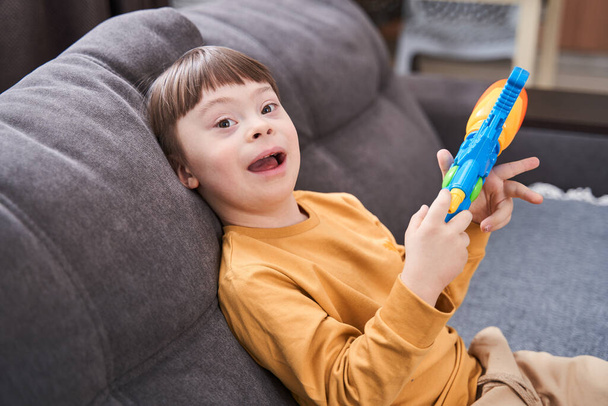 Portrait view of the overjoyed boy with genetic disorder sitting at the sofa and laughing while playing with toy gun. Happy childhood concept - Фото, изображение