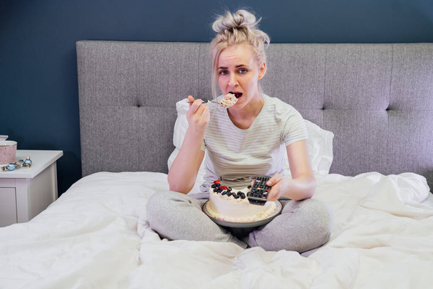 Bored, sad woman in home clothes eating cake and using the remote control to change the channel while watching television in bed at night. Overeating, Jamming of negative emotions. Selective focus - Фото, изображение