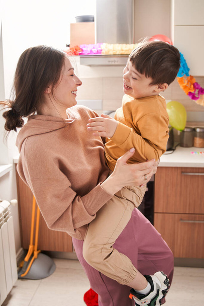Vertical view of the overjoyed woman holding at the hands her male child with genetic disorder while having fun at the kitchen - Фото, изображение
