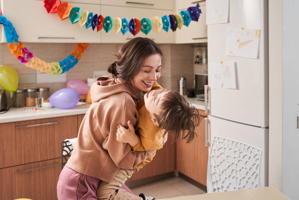 Cute little boy with Down syndrome embracing his lovely mother while she holding him at the hands at the decorated kitchen - Фото, изображение