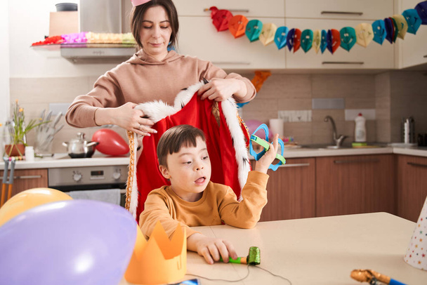 Friendly caucasian woman wearing paper crown preparing celebrating birthday with her lovely son with genetic disorder while putting king costume at him - Foto, Imagem