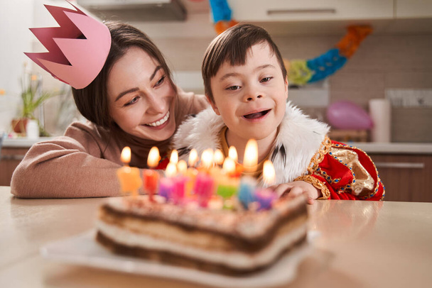 Cute boy with Down syndrome looking with interest at the cake with candles while celebrating his birthday with mother at home - Foto, afbeelding