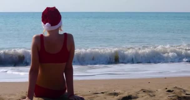  Sırtı parlak Noel Baba şapkasıyla mutlu bir kız deniz kıyısında güneşe karşı oturur. Yeni yıl tatili - Video, Çekim