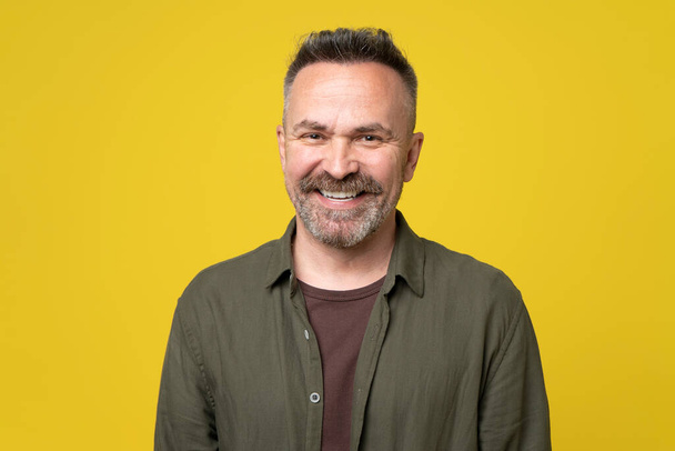 Portrait of 50s handsome smiling man with beard and short haircut wearing casual green shirt isolated over yellow background. Attractive happy man is smiling. Positive middle-aged active person. - Photo, Image