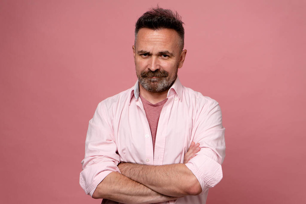 Mature handsome man with beard and mustache dressed in pink pastel shirt with crossed hands isolated over pastel red background frowning face looking at camera. Positive cheerful middle-age person. - Фото, зображення