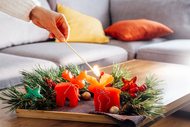Female hand lighting a candle with a match in Christmas cozy composition for hygge home mood. Deer shape red candles, fresh green pine branches and Xmas decor on the coffee table. Selective focus. - Fotoğraf, Görsel