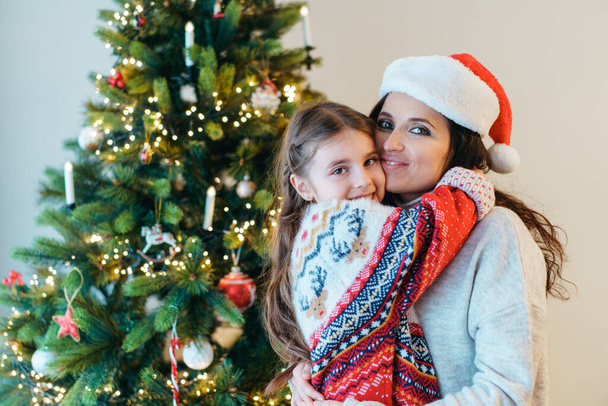 Portraits of smiling little daughter hugging her smiling mother in santa hat on Christmas tree background. Family holidays. Festive christmas mood. Happy childhood. Selective focus. - Фото, зображення