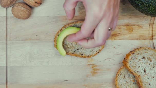 vrouwelijke hand zet stukken avocado op brood, op een houten plank. - Video