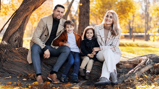 Bonne famille dans un parc d'automne. Mère, père, fils et fille sont assis sur un tronc d'arbre et regardant dans la caméra, arbres jaunis autour - Photo, image
