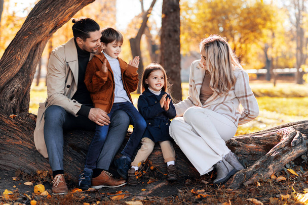 Ευτυχισμένη οικογένεια σε ένα φθινοπωρινό πάρκο. Μητέρα, πατέρας, γιος και κόρη κάθονται σε ένα κορμό δέντρου, κιτρινισμένα δέντρα γύρω - Φωτογραφία, εικόνα