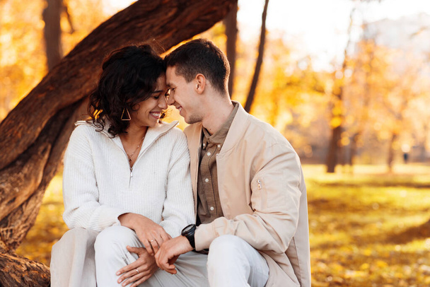 Una coppia romantica in un parco autunnale. Seduto su un tronco d'albero e sorridente. Atmosfera autunnale, alberi ingialliti e foglie intorno - Foto, immagini