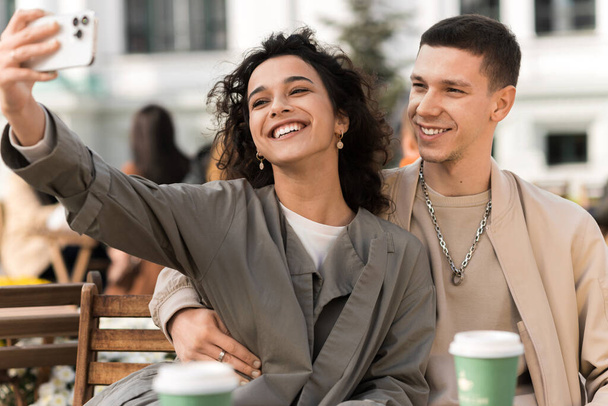 Ein glückliches Paar im Freien in der Nähe eines Cafés. Selfie machen, lächeln, Kaffee auf dem Tisch. Herbststimmung - Foto, Bild