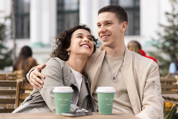 Una pareja feliz al aire libre cerca de un café. Abrazándose, sonriendo, tomando café en la mesa. Ambiente otoñal - Foto, Imagen
