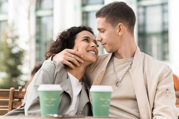 Una pareja feliz al aire libre cerca de un café. Abrazándose y mirándose el uno al otro, café en la mesa. Ambiente otoñal - Foto, Imagen