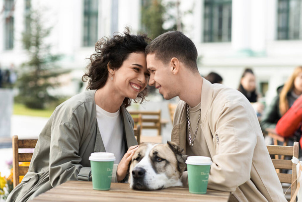 Счастливая пара на улице возле кафе. Улыбаются, гладят собаку, пьют кофе на столе. Осенняя атмосфера - Фото, изображение