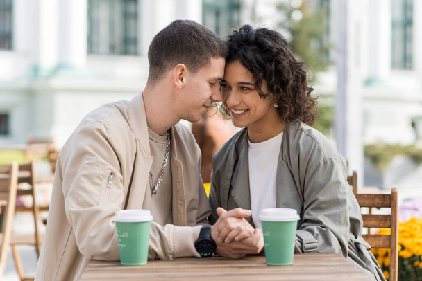 Um casal romântico ao ar livre perto de um café. De mãos dadas, a sorrir, café na mesa. Ambiente de outono - Foto, Imagem