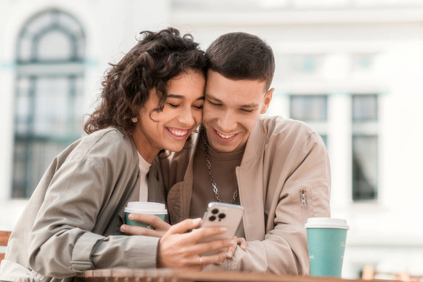 Una pareja feliz al aire libre cerca de un café. Mirando en el teléfono, sonriendo, hablando, café en la mesa. Ambiente otoñal - Foto, imagen
