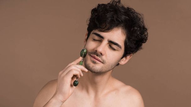 A young caucasian man with stubble beard is doing a facial massage using a roller - Photo, Image