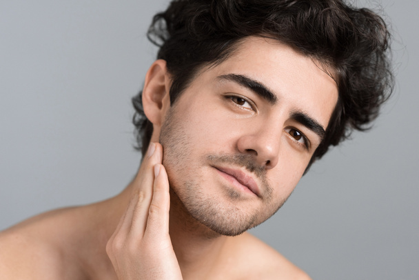 Egy fiatal, borostás szakállú kaukázusi férfi arc- és bőrápolást végez kozmetikai olajjal arcmasszázs közben, belenéz a kamerába. - Fotó, kép