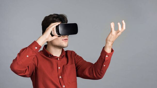 Ein junger Mann spielt mit einer VR-Brille, hält ein virtuelles Ding, grauer Hintergrund - Foto, Bild