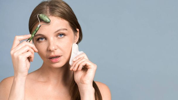 Young caucasian woman posing with roller and Gua Sha for facial massage, looking into the camera - Photo, Image