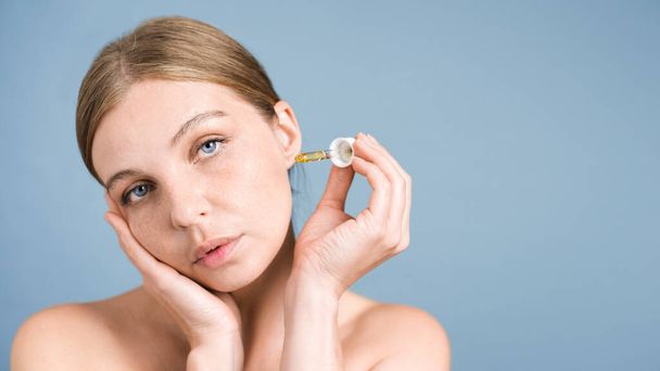 Uma jovem caucasiana está aplicando e hidratando rosto e pele com óleo cosmético, olhando para a câmera - Foto, Imagem
