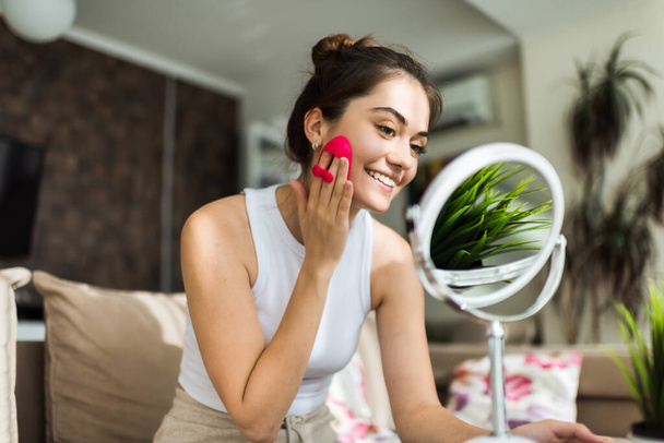 Uma jovem caucasiana sorrindo mulher está fazendo uma massagem facial olhando no espelho - Foto, Imagem