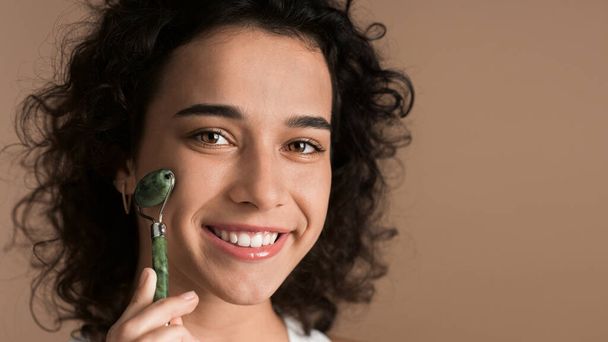 Uma jovem mulher branca sorrindo está fazendo uma massagem facial usando um rolo, olhando para a câmera - Foto, Imagem