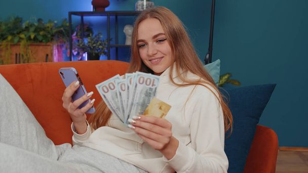 Planning family budget. Smiling lovely girl counting money dollar cash, use smartphone calculate domestic bills at home. Joyful young woman satisfied of income saves money for planned vacation, gifts - Foto, Imagem