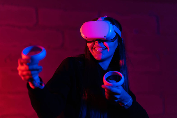 Młoda podekscytowana kobieta grająca w gry VR z okularami i kontrolerami VR. Oświetlenie czerwone i niebieskie - Zdjęcie, obraz
