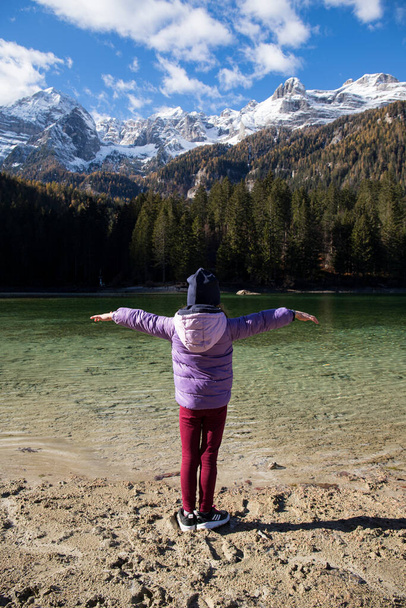 Дитяча дівчинка стоїть з розкритими обіймами біля кришталево чистих вод озера Товель, Трентіно Альто Адіж, Італія. Осінній вид з сніжними горами на задньому плані. - Фото, зображення