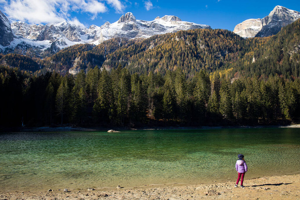 Niña de pie cerca de las aguas cristalinas del lago Tovel, Trentino Alto Adigio, Italia. Vista de otoño con montañas nevadas en el fondo. - Foto, Imagen