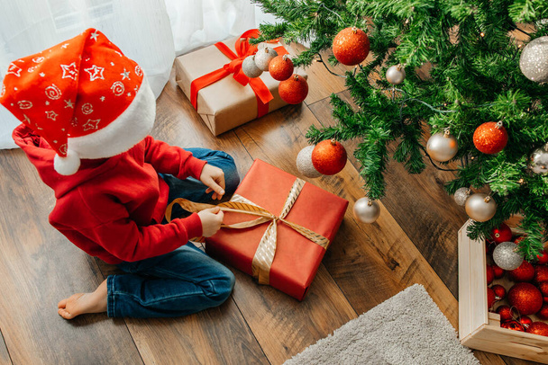 Nowy Rok i Boże Narodzenie. Chłopiec rozwiązuje łuk i otwiera pudełko prezentem świątecznym.. - Zdjęcie, obraz