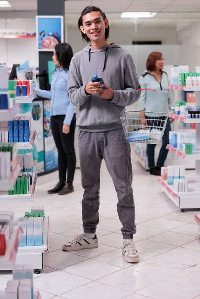 Asian male client coming at pharmacy shop to buy medication and prescription treatment, looking at boxes of health care products on shelves. Customer holding pharmaceutical supplies in drugstore. - Photo, Image