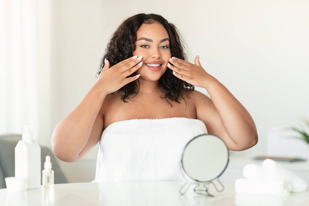 Genç Afrikalı Amerikalı artı cüsseli kadın yatak odasında yüz bakımı yaparken yüzüne dokunuyor cilt durumunun keyfini çıkarıyor ve kameraya gülümsüyor. - Fotoğraf, Görsel