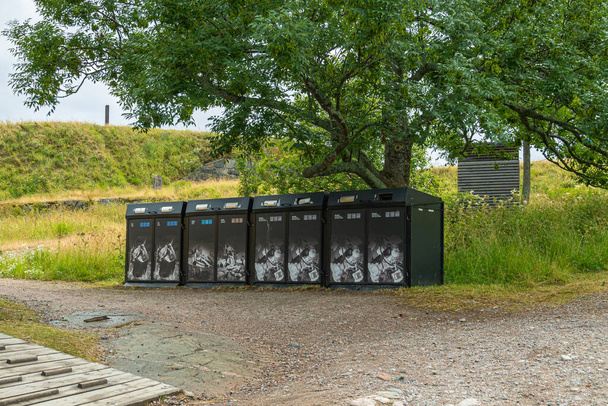Helsinki - 19. heinäkuuta 2022: Suomenlinnan linnoitus. Rivi mustia roskakoreja asetettu pitkin soraa polku alla vihreä lehvistö puu - Valokuva, kuva