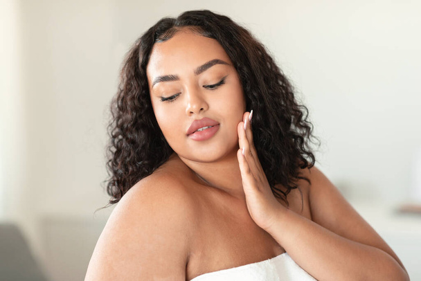 Attractive african american plus size woman touching her face and enjoying silky touch after using brand new nourishing cream, bedroom interior, copy space - Valokuva, kuva