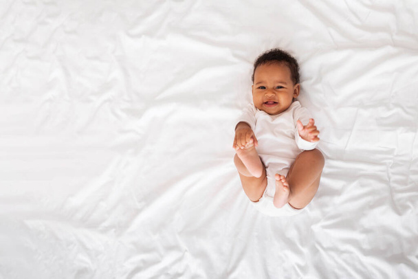 Funny Little African American Baby Resting On Bed In Bedroom, Cute Black Infant Boy Playing With His Toes And Looking At Camera While Relaxing At Home On White Linens, Top View With Copy Space - Photo, Image