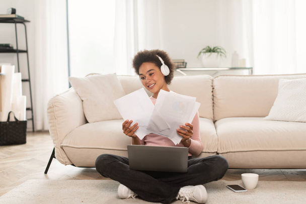 Happy millennial african american female in wireless headphones has video call on laptop, works with documents sits on floor in bright living room interior. Paperwork, business, study remotely at home - Photo, Image