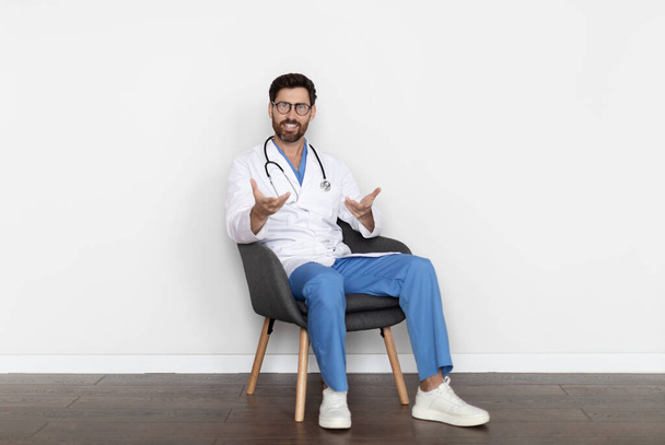 Όμορφος μεσήλικας άνδρας γιατρός με στολή που κάθεται στην καρέκλα και μιλάει στην κάμερα, σίγουρος γιατρός άντρας με λευκό παλτό και στηθοσκόπιο σύμβουλος ασθενή, δίνοντας ιατρική συμβουλή - Φωτογραφία, εικόνα