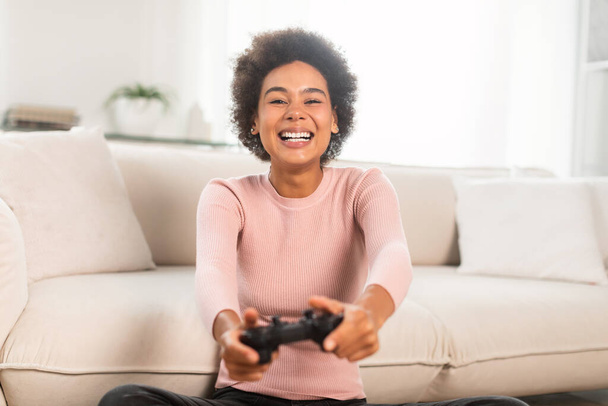 Happy excited millennial african american female with joystick, sit on floor, playing online games in light living room interior. Entertainment with device in spare time at home alone and emotions - Foto, Imagen