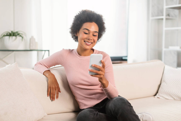 Hymyilevä melko tuhatvuotinen afrikkalainen amerikkalainen nainen chattailuun älypuhelimella, istua sohvalla valoisa olohuone sisustus. Lady katsella video, lukea blogia, surffaus internetissä ja sosiaaliset verkostot yksin kotona - Valokuva, kuva