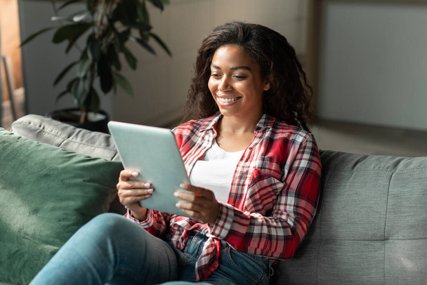 Весёлая чёрная молодая женщина читает блог, чатится на планшете, смотрит видео, наслаждается онлайн-шоппингом в интерьере гостиной. Новое приложение, социальные сети для общения, крепостное право и новая норма жизни дома - Фото, изображение