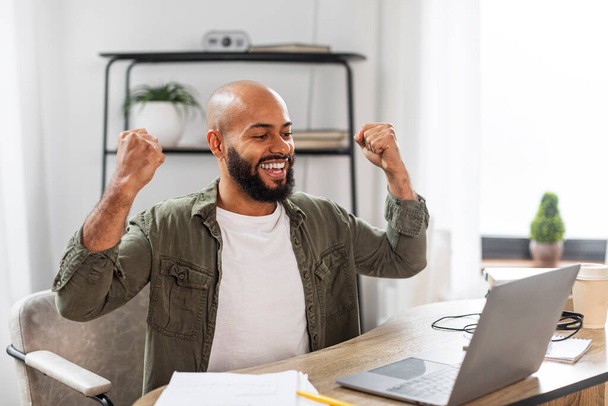Vreugdevolle volwassen mannelijke ondernemer met behulp van laptop en gebaren ja, vieren zakelijk succes tijdens het werken thuis. Emotionele freelancer man schudden vuisten in vreugde - Foto, afbeelding