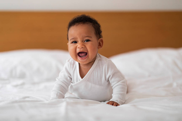 Adorable Small Black Baby Crawling On Bed And Laughing, Portrait Of Cute Little African American Infant Boy Or Girl Lying On His Tummy, Relaxing On White Bedsheets In Bedroom At Home, Copy Space - 写真・画像