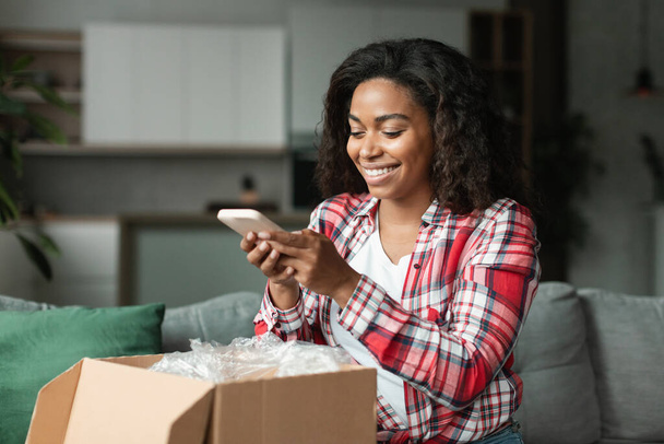 Happy pretty black young woman client in casual unpacks parcel, takes photo of purchases on smartphone in living room interior. Fashion blog and online shopping, gadget for reviews on buy and delivery - Foto, Imagen